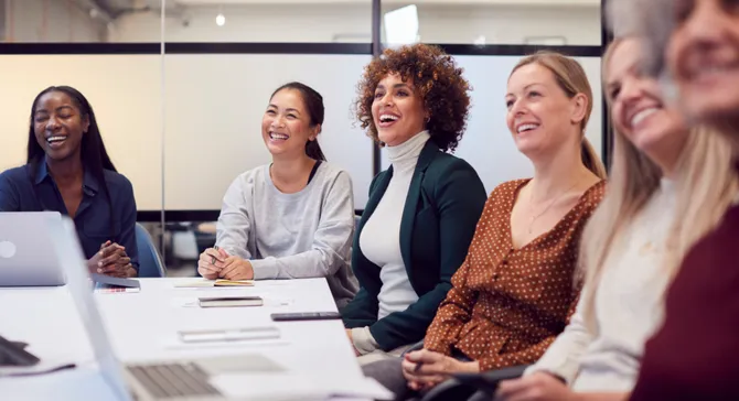 A group of women in smart casual dress sat around a table in a meeting room, smiling.