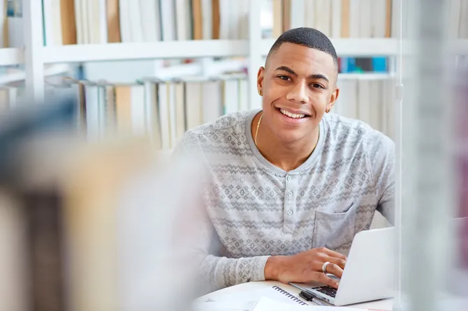 A person sitting at a desk in Newport library with their laptop, smiling at the camera. There are bookshelves in the background.