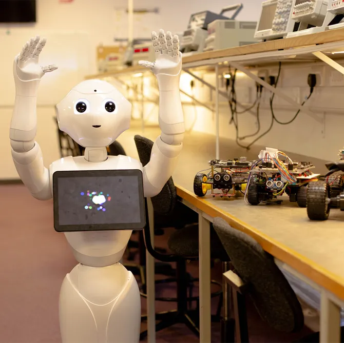 Pepper the robot holds its hands up in the air while being stood in front of electric cars in a USW Treforest engineering workshop