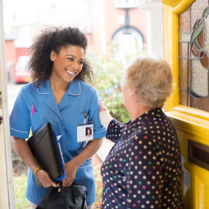 A female nurse or community care worker is stepping through the front door, with a residential street in the background, being greeted by a senior female patient. She is wearing a blue nurses tunic , and holding a medicines bag. She is wearing an ID badge with her profile photo already on it.