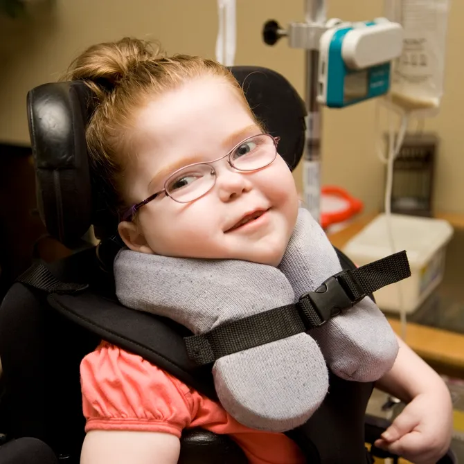 Little girl with cerebral palsy smiles at camera while sitting in her wheelchair at home with other medical equipment in the background