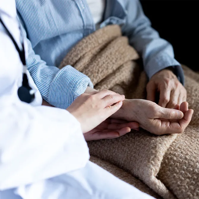 A close up on the hand of a medical professional, wearing a white laboratory coat and a stethoscope, resting their hand on an elderly patient, wearing pyjamas with a blanket on their lap, to comfort them while talking