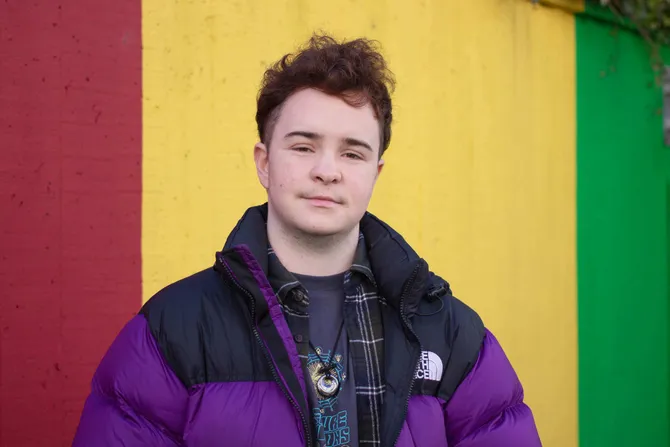 A profile of law student, Ash Taylor, stood against a red, yellow and green painted wall.
