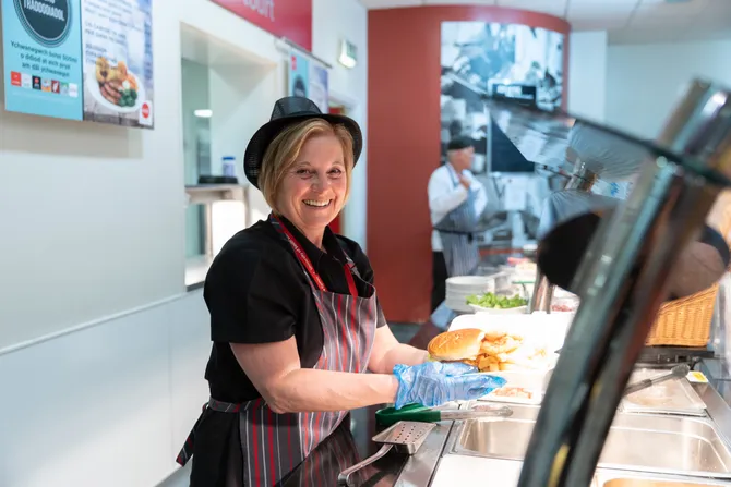 a member of University catering staff, standing behind a hot food counter serving a burger and chips