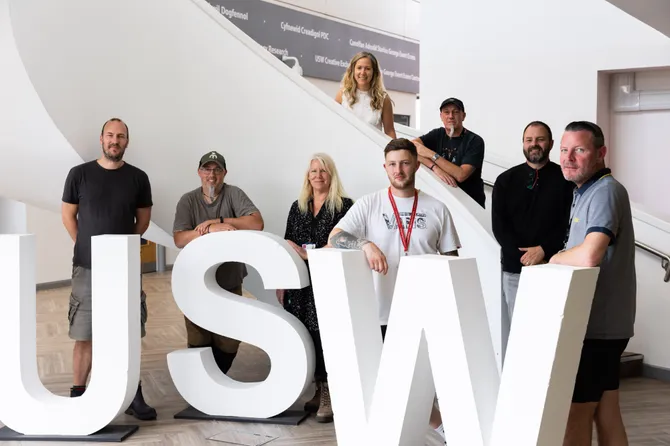 8 members of staff standing on the spiral staircase at the Cardiff Campus with a USW sign, smiling at the camera