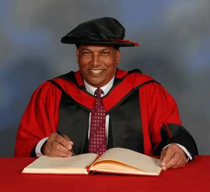 Raj Aggarwal smiling at the camera while wearing a doctorate's gown and cap and writing in a book on a red desk in front of a blue backdrop