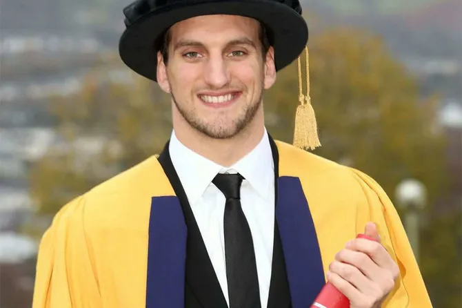 Sam Warburton smiles at the camera and holds their degree outside while wearing a doctorate cap and gown