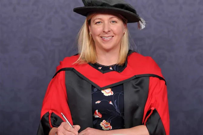 Jayne Ludlow smiles at the camera while wearing a doctorate's cap and gown in front of a blue backdrop