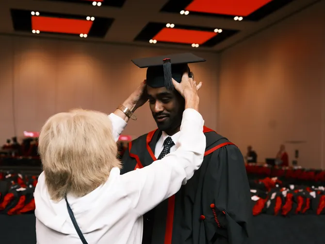 A member of staff in a white shirt helps a student to position their graduation cap at the ICC in Newport in the robing room