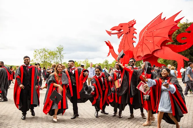 A group of students in graduation gowns throwing caps into the air in front of the Welsh dragon.