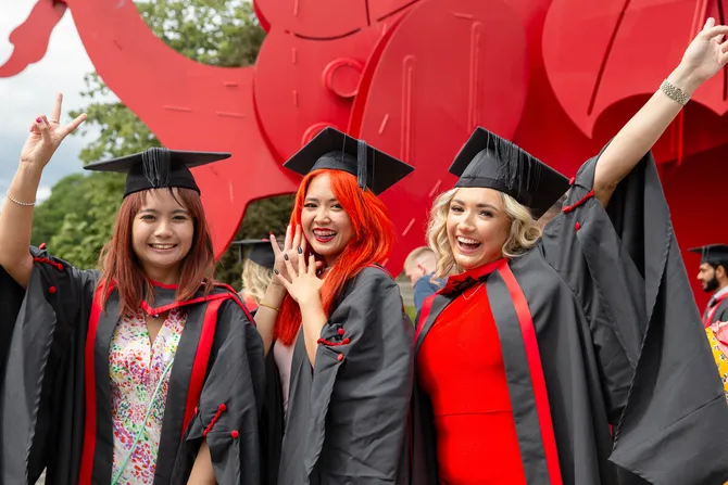 Three students dressed in graduation robes and graduation caps smile and pose in front of the red dragon outside Newport's ICC