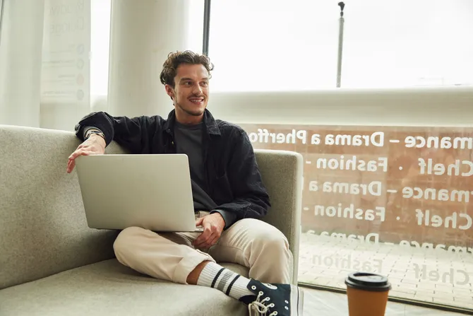 A student sat on a sofa in front of a window with a laptop.