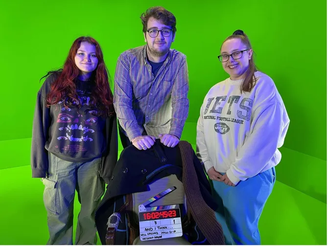 Three students are stood, smiling, in front of a green screen with a clapboard in front of them.