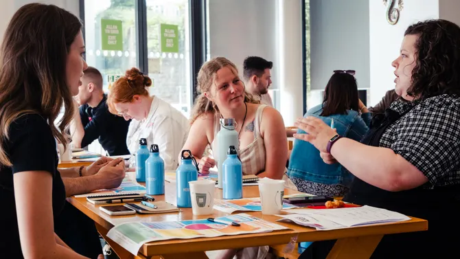 A group of students in conversation around a table, covered in worksheets and notepads
