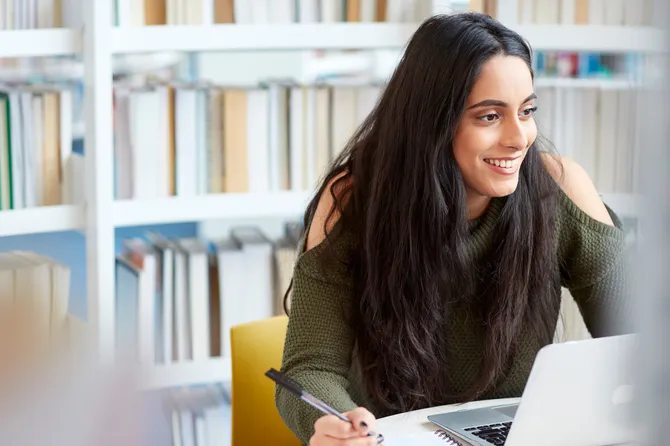 A student is sat smiling in a library, they are working on a computer.
