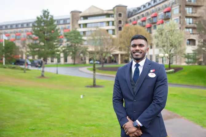 A student in hotel uniform is smiling into the camera. They are standing outside the Celtic Manor Resort.