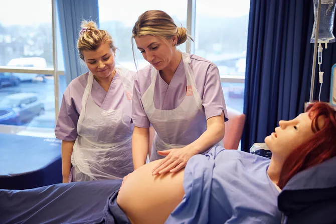 A Midwifery student is feeling the belly of a pregnant dummy, while being supervised.