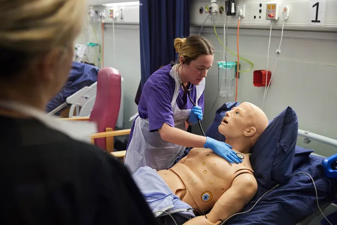 An academic is supervising a nursing student listening to the heart of a dummy patient.