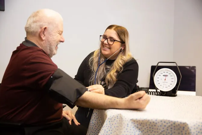 An older man is having his blood pressure checked by a healthcare worker in what looks like a clinic or medical office. The healthcare worker is smiling while using a stethoscope and a blood pressure cuff wrapped around the man’s arm. A blood pressure gauge sits on the table nearby, and the interaction appears friendly and relaxed.