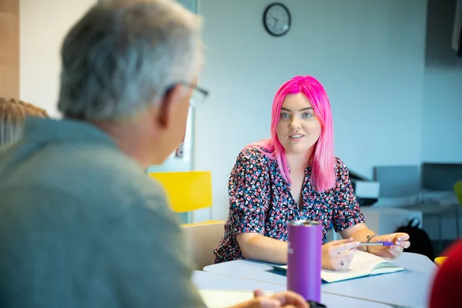 A student with shoulder-length bright pink hair listens intently to a fellow student while taking notes in a practice counselling session
