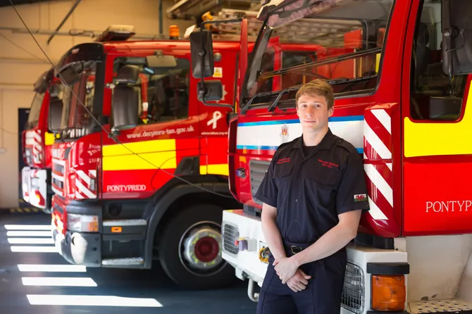 A student in uniform is standing with arms folded in front of two fire engines.