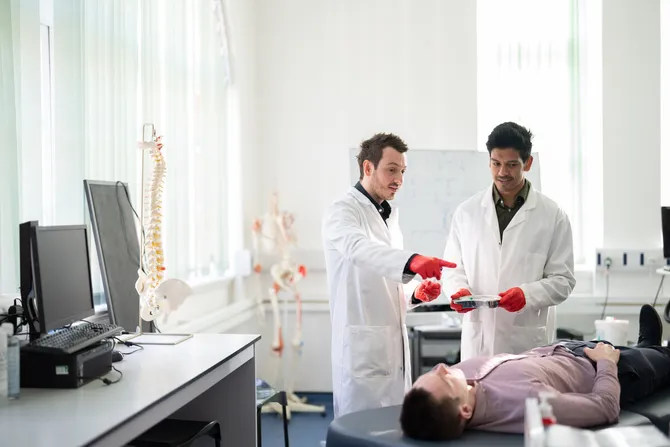 Two students in a sports science lab in conversation whilst a patient lies on a bed in front of them