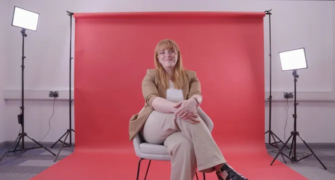 university staff member sitting on a chair in front of a red background smiling to camera