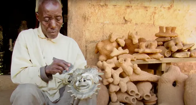 A man is sat on the floor outside of a beige stone building adding finishing touches to a metal sculpture.