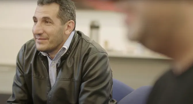 A man wearing a blue shirt and a leather-style jacket is sat down looking at someone with a soft smile.