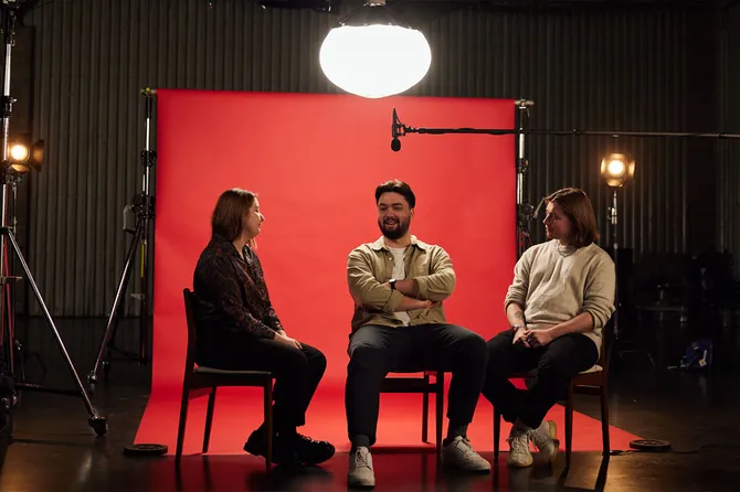 Graduates Savva, Sam and Niamh, sat together on chairs chatting in front of red backdrop