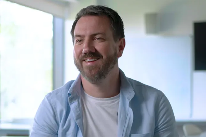 A man in a white tshirt and blue overshirt is smiling on camera.