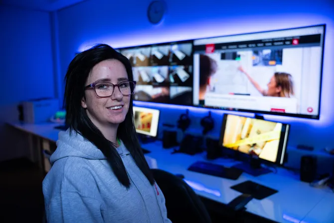 A student smiles at the camera in a dim dark blue room in front of four computer monitors showing surveillance footage and the USW website