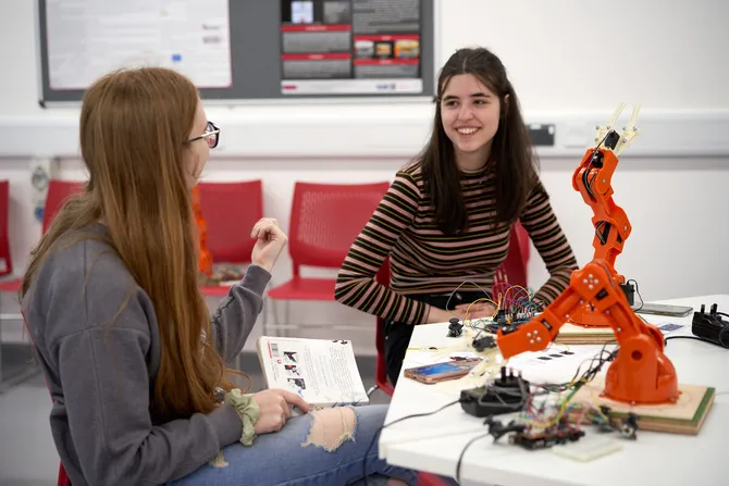Two school students smile while chatting with books in an engineering workshop near a robotic arm