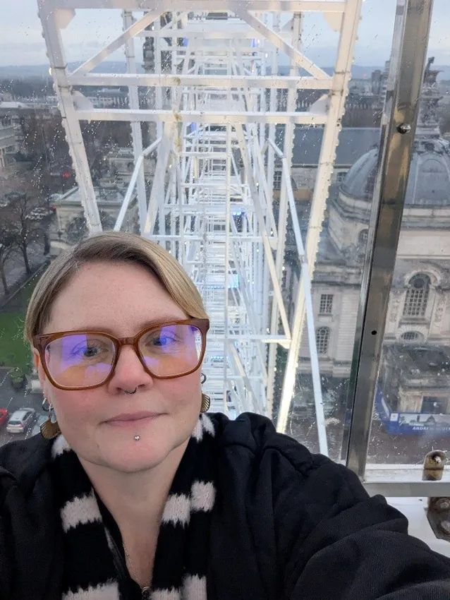 Laura Roberts ar olwyn fawr gyda’r strwythur gwyn y tu ôl a golygfa o’r ddinas lawog oddi tano.m and a rainy city view below