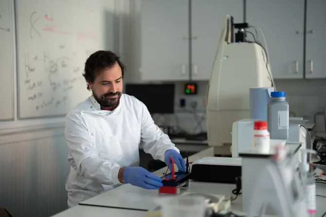 A postgraduate student wearing a white laboratory coat and blue medical gloves conducts a wildlife ecology experiment using a small electrical device in a biological sciences laboratory