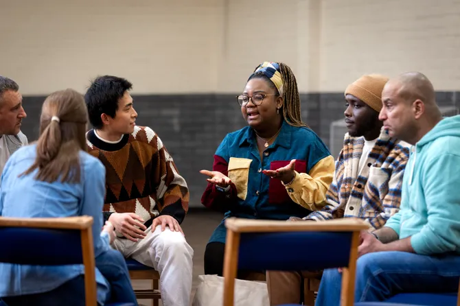A group of adults dressed in casual clothes sitting in a circle and talking to each other about their mental well-being.