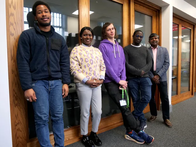 A group of five students of various ages smile while standing in front of a meeting room in the Postgraduate Centre at the USW Treforest campus