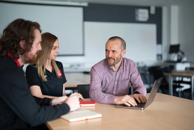 A group of three sport researchers, including PhD students and doctoral supervisors, sit around a table chatting with notepads and a laptop