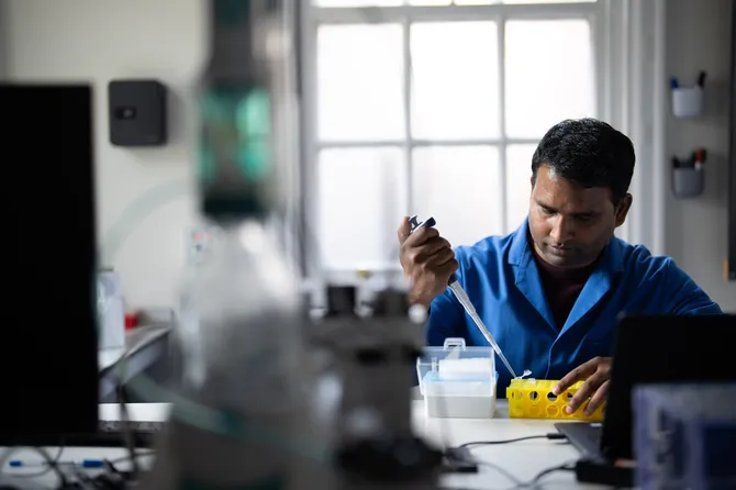 A student in blue overalls uses a pipet to transfer liquid into a test tube in a yellow test tube holder in a laboratory surrounded by scientific equipment
