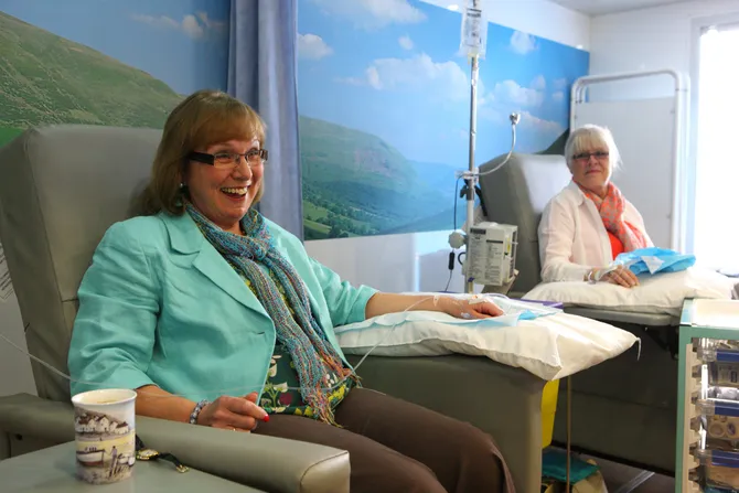 Two patients sit in chairs with their arms supported by pillows while attached to medical machines administering treatment in a hospital ward