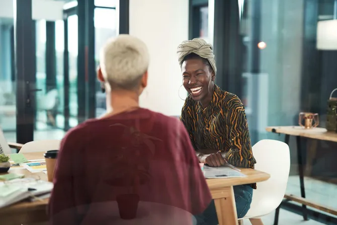 Two women sat at a desk filled with papers smiling and laughing.