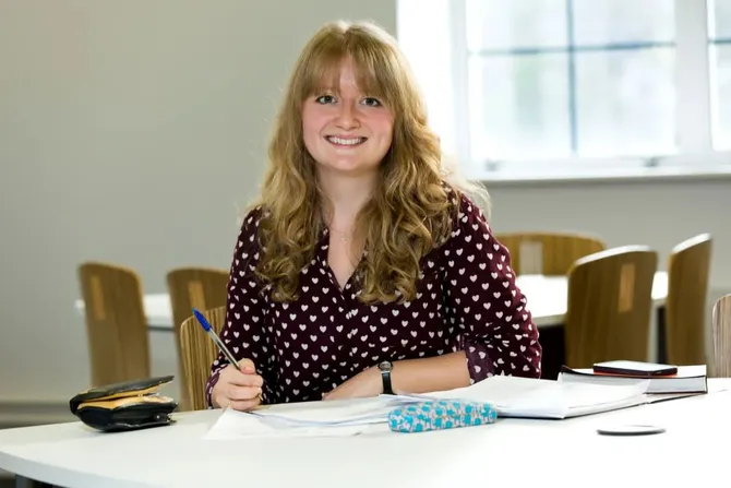 Sophie John, PhD student in Health Sciences and Psychology, sat at a table surrounded by pieces of paper, smiling at camera