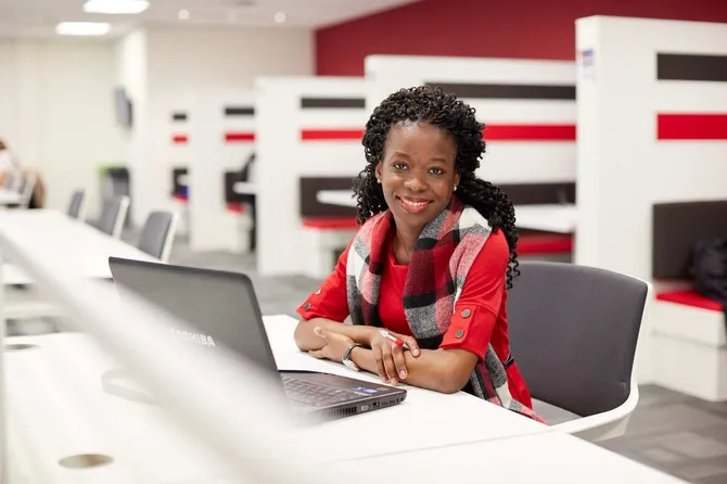 A student sat in the library working on a laptop and smiling at the camera.