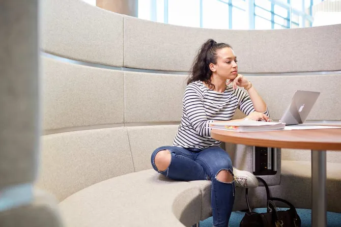 A student sitting on a sofa in a study area looking at a laptop on a table.