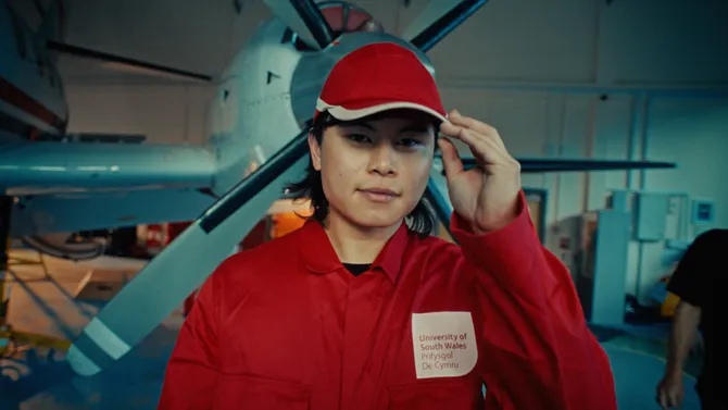 Close up shot of an engineering student, stood in front of a plane in the aerospace centre, wearing red USW overalls