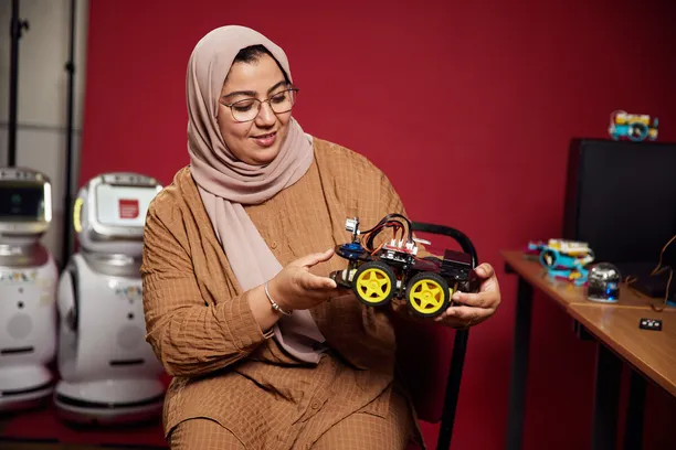 Senior Lecturer, Dr Mabrouka Abuhmida, holding a robotic car model in front of a red backdrop, with two small robots in the background