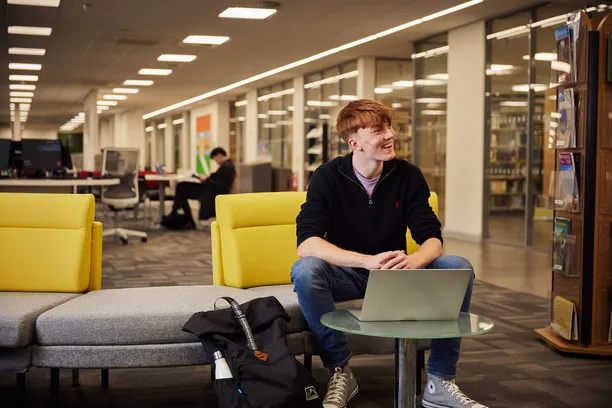 A student smiling in the library with books and study spaces in the background.