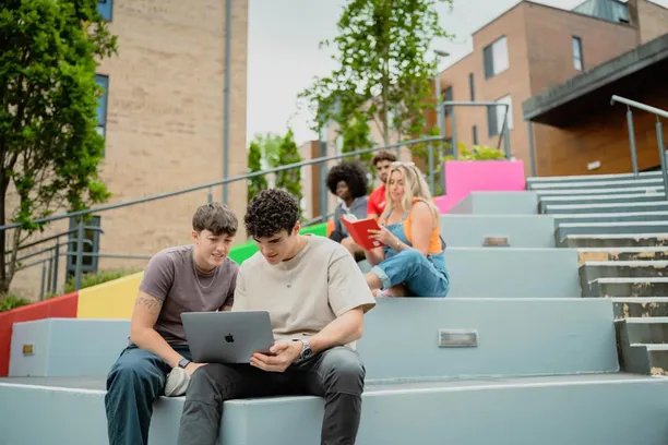 Two students are sat on colourful steps, looking at a laptop, outside the accommodation hub, with another reading a book behind them.