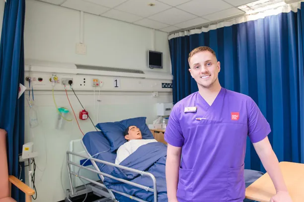 Nursing student, Aston, is stood at the end of a hospital bed in the Clinical Simulation Centre. He is wearing purple USW scrubs.