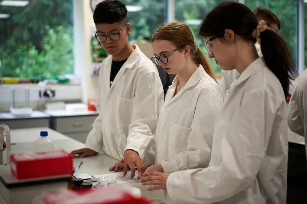 Three school pupils dressed in white laboratory coats discuss an ongoing experiment and their notes in a science workshop